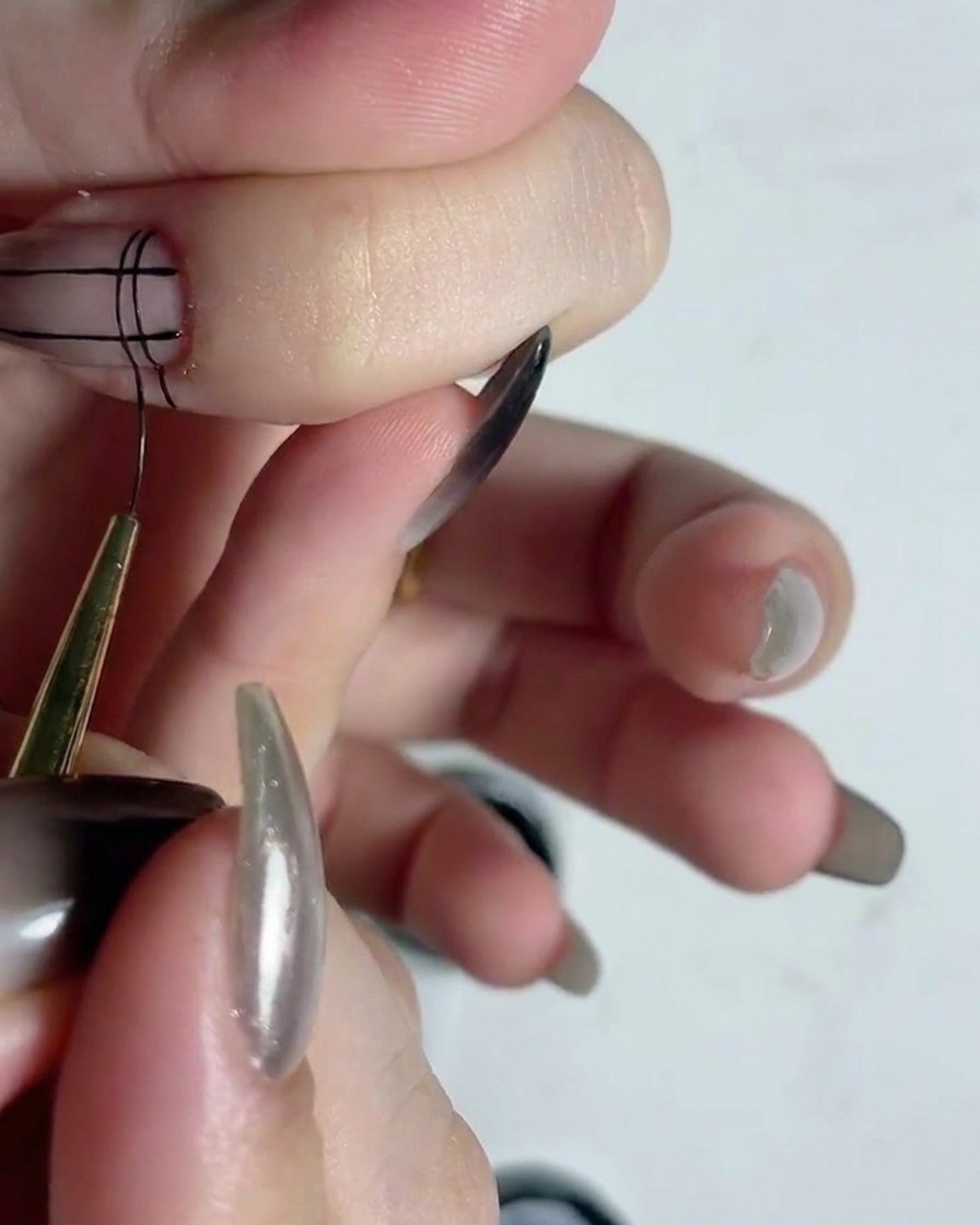 Ein feiner Liner-Pinsel zeichnet horizontale schwarze Linien über vertikale Linien, um ein Gittermuster auf einem Fingernagel zu erzeugen.