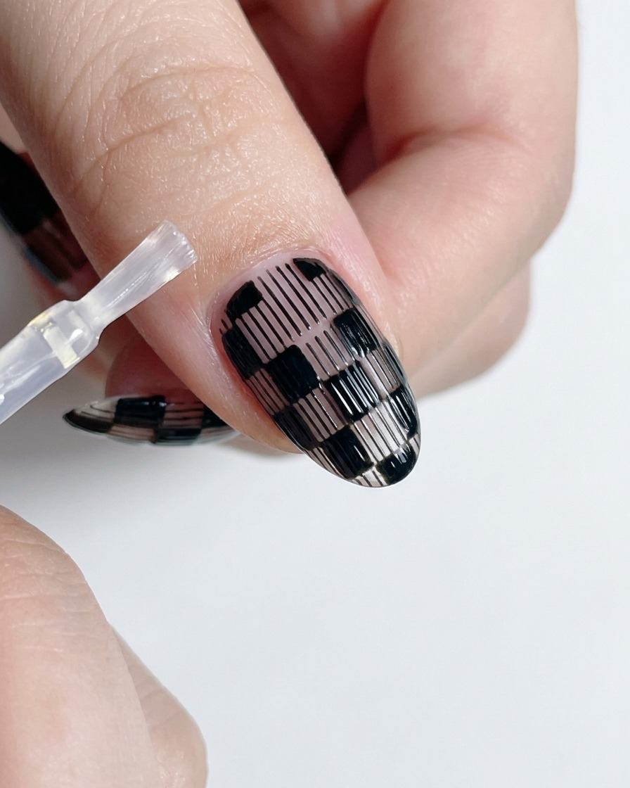Ein Top-Coat-Pinsel trägt klaren Glanzlack über ein fertiges schwarz-weißes Karo-Nageldesign auf.
