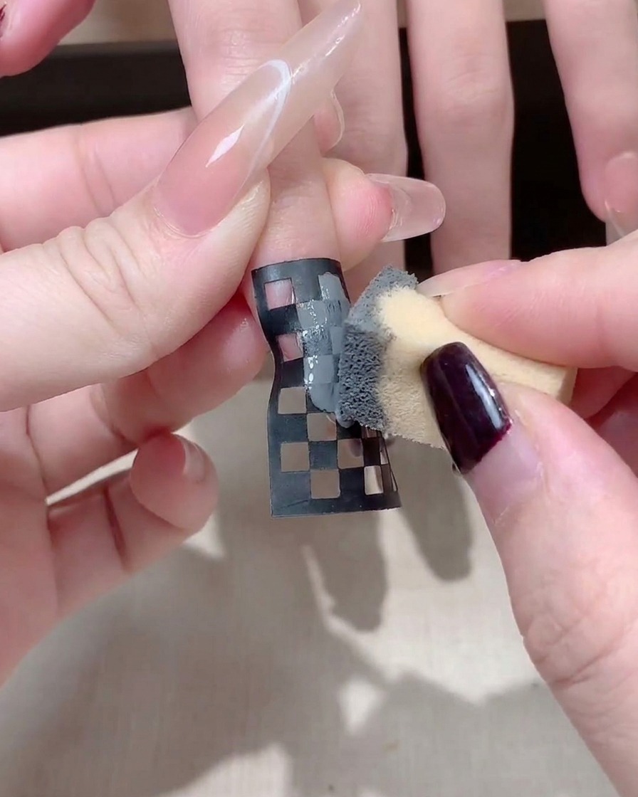 Nahaufnahme einer Nageldesignerin, die einen schwarz-weißen Schachbrett-Nagelsticker auf einem langen quadratischen Nagel anpasst.