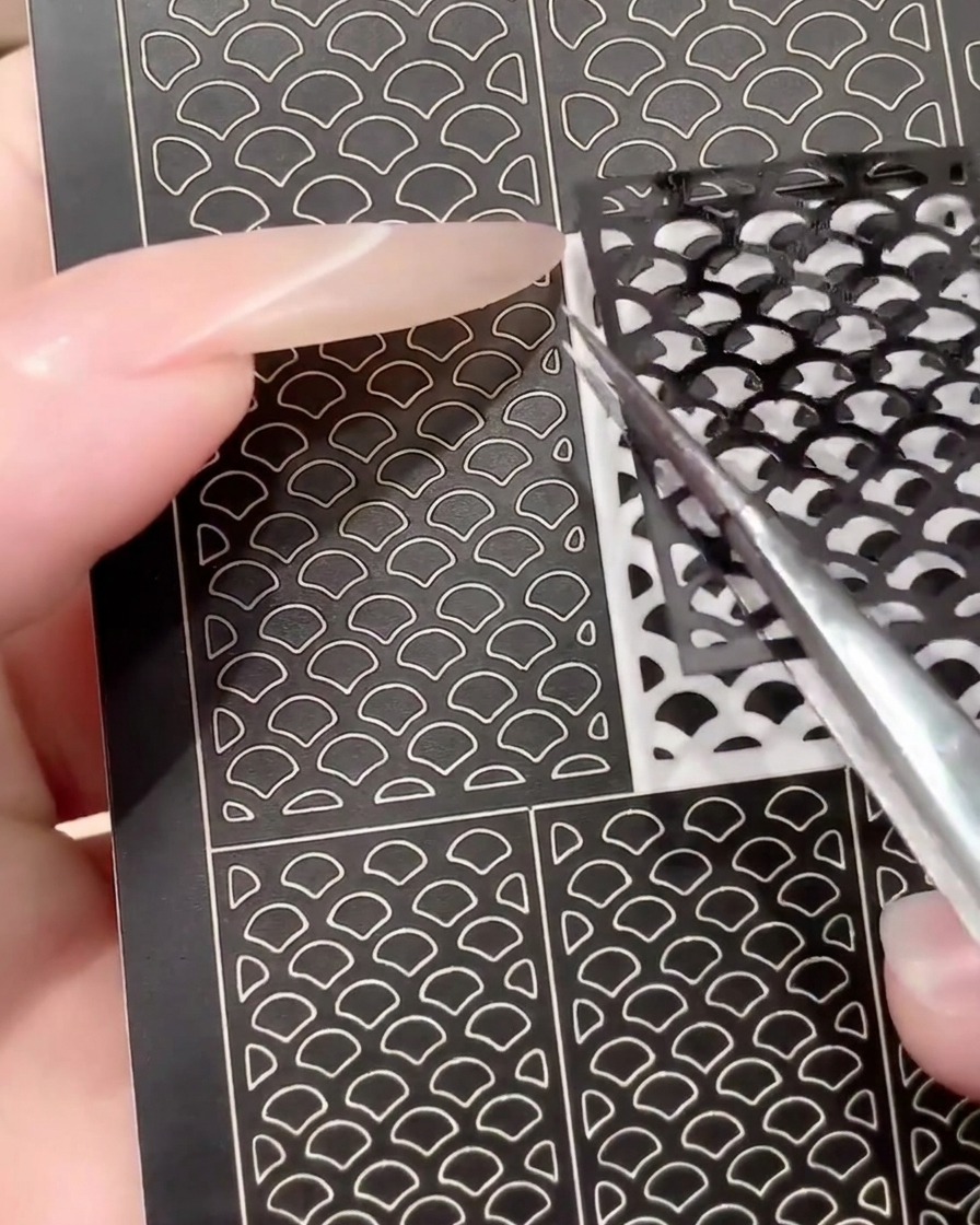 Eine Pinzette zieht einen benutzten, schwarzen Nail-Art-Stickerbogen ab, um die Wiederverwendbarkeit zu demonstrieren.