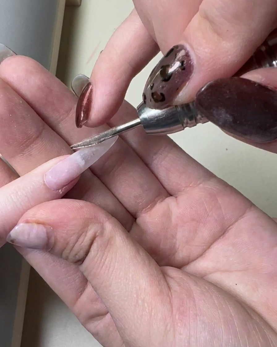 Ein Nagelpinsel trägt eine dünne Schicht transparentes, graues Gel auf einen langen, vorbereiteten Kunstnagel auf.