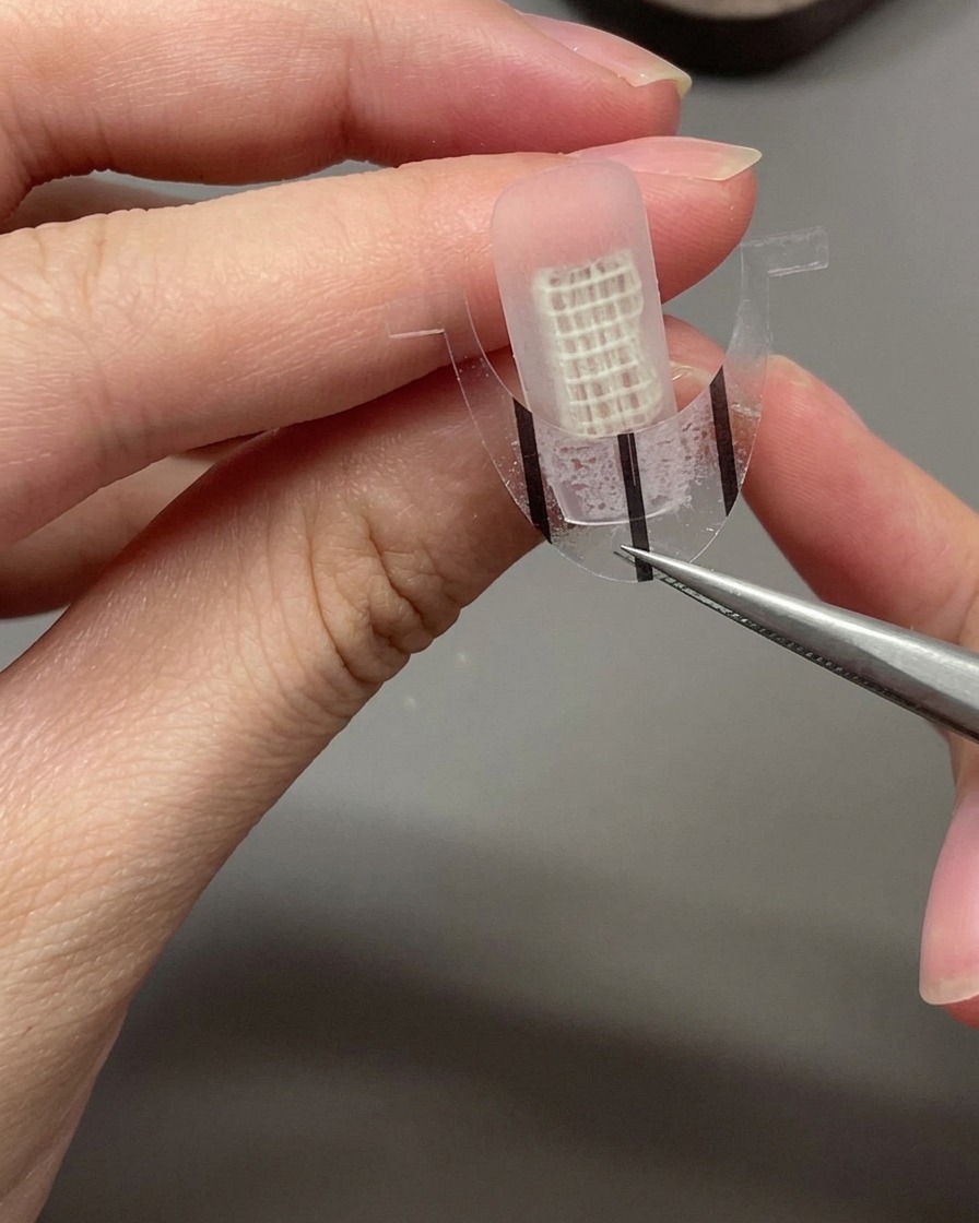Eine Pinzette platziert vorsichtig einen gebogenen French Hilfsaufkleber über die Mitte eines unlackierten Nagels.
