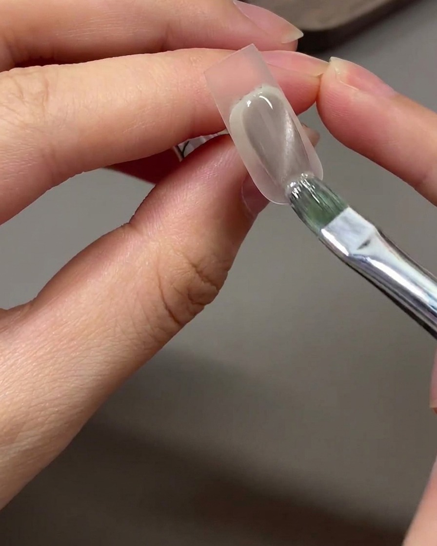 Ein Pinsel trägt grau-nudes Cat Eye Gel-Polish auf den mit dem Schwämmchen getupften Spitzenbereich eines Nagels auf.