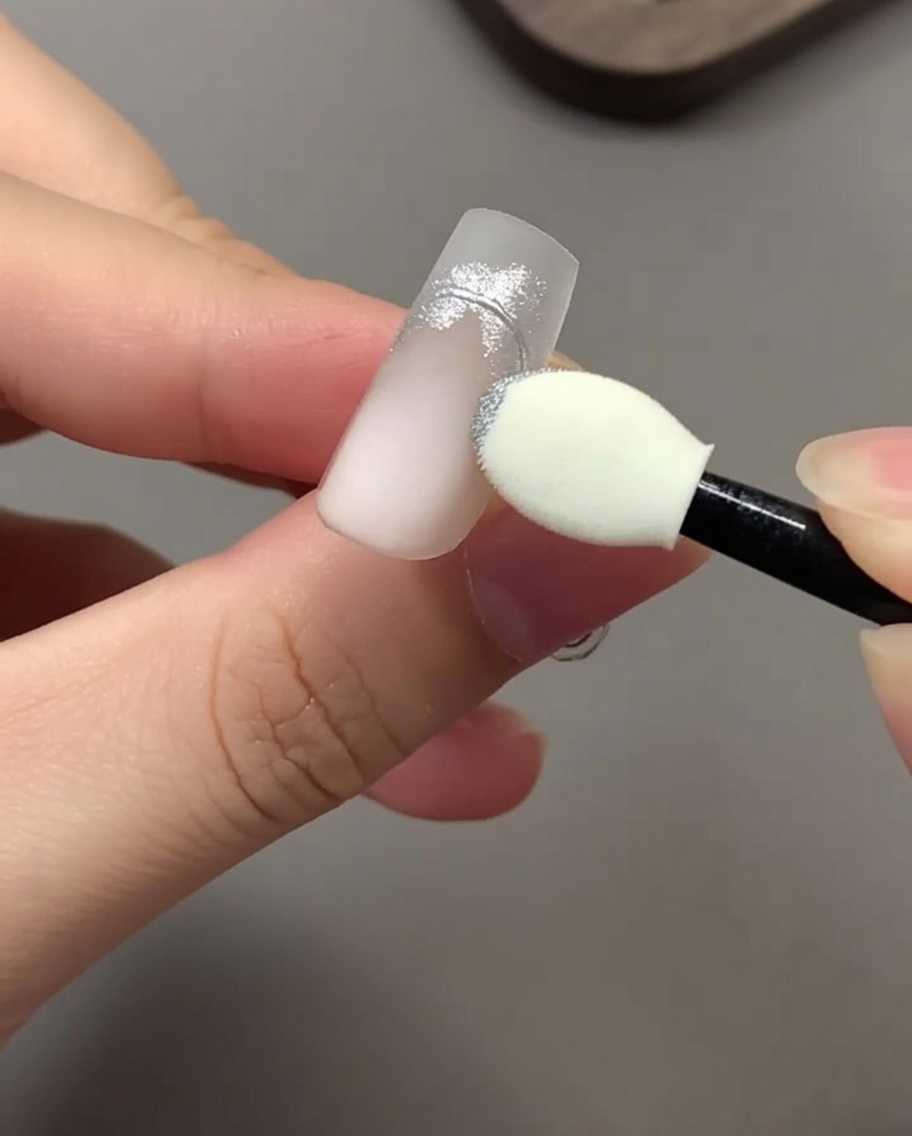 Ein Lidschatten-Schwammapplikator reibt silbernes Chrom-Puder auf ein Nageldesign.