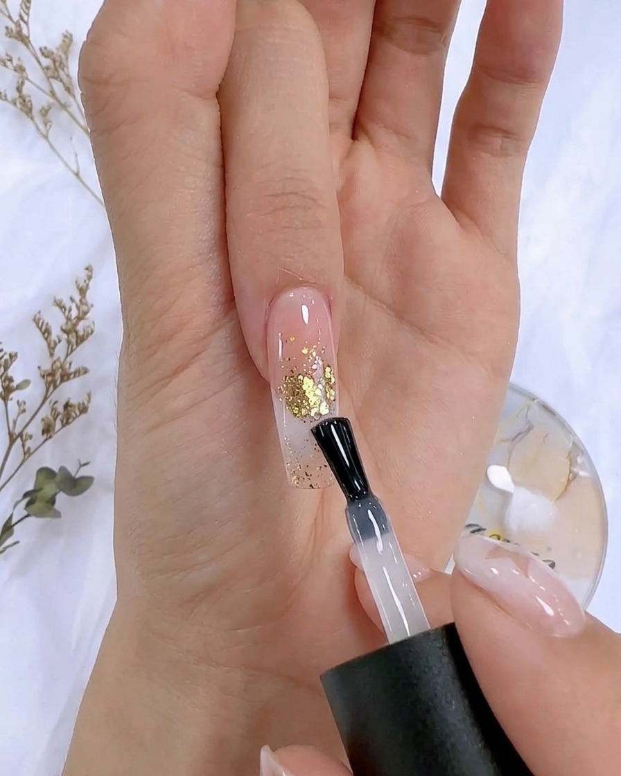 Ein Nagellackpinsel trägt einen klaren Top-Coat auf einen dekorierten Nagel mit Blattgold-Akzenten auf.