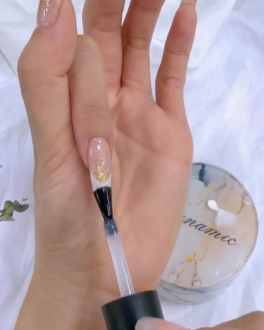 Eine Hand hält eine Top-Coat-Flasche während des finalen Aushärtungsschritts in die Nähe einer UV/LED-Nagellampe.
