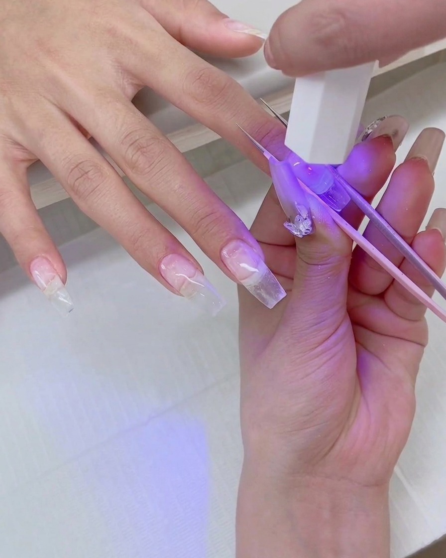 Nahaufnahme einer Hand, die eine Mini-UV-Blitzlampe über einen klaren Nagel-Tip hält, während eine Pinzette den Tip fixiert.