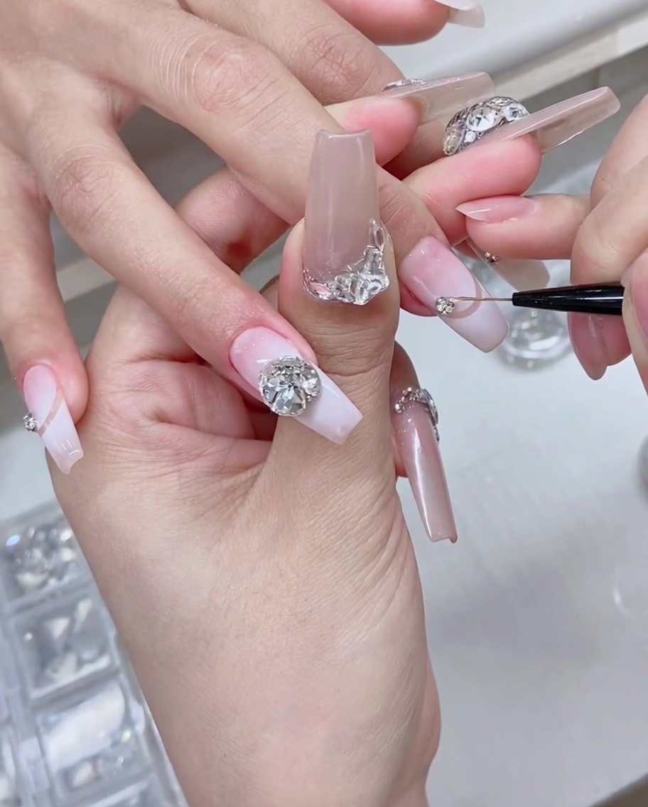 Ein feiner Detailpinsel und eine Pinzette platzieren funkelnde silberne Strasssteine auf der Basis eines manikürten Nagels.