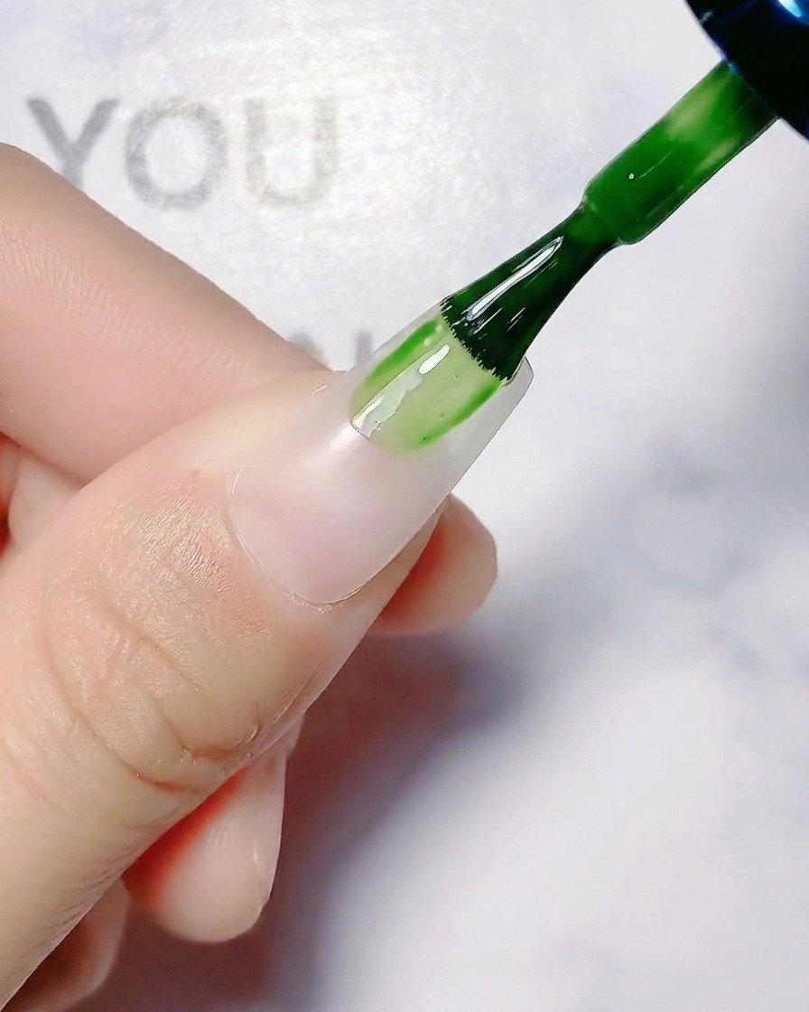 Ein Nagelpinsel trägt transluzenten weißen Basislack auf einen unlackierten Fingernagel auf.