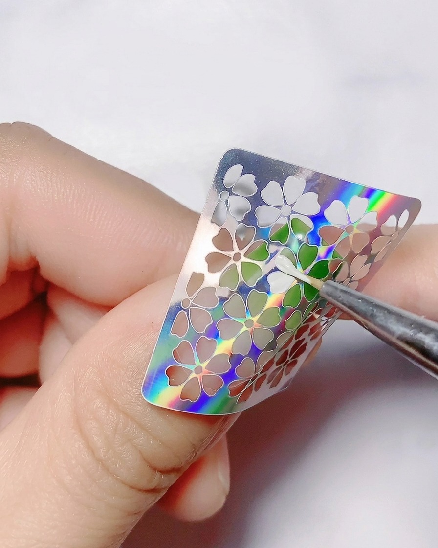 Ein feiner Pinsel trägt weißes Farbgel in die hohlen Blumenausschnitte eines Stickers auf.