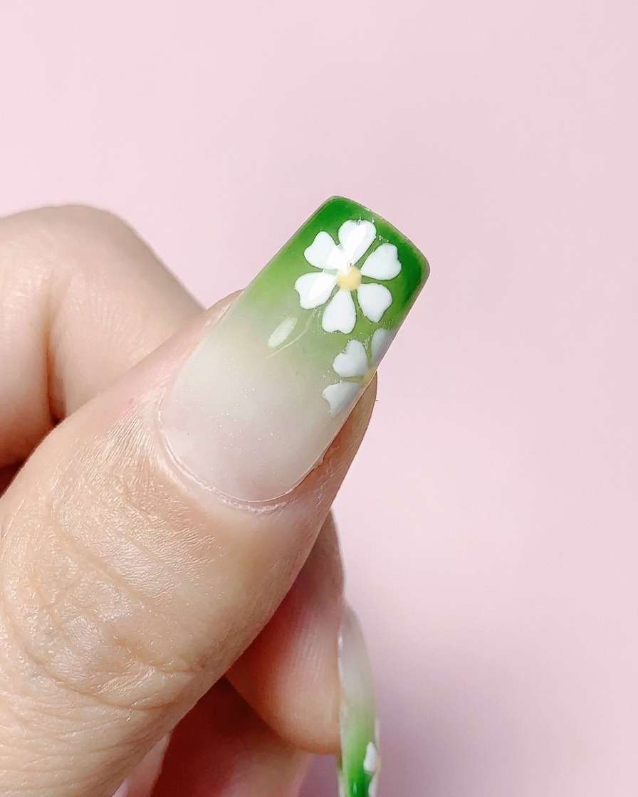 Ein fertiger quadratischer Nagel mit grünem Farbverlauf und weißen Blumen wird mit einem klaren Topcoat überzogen.