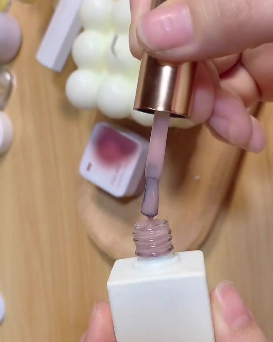 Ein Pinsel zieht transluzenten Rosa-Unterlack aus einer weißen Nagellackflasche heraus.