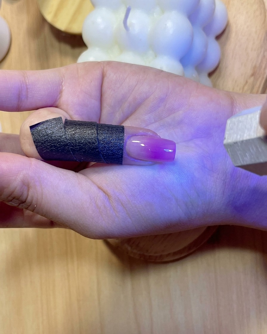 Ein Fingernagel glimmt unter violettem UV/LED-Licht beim Aushärten des aufgetragenen Verstärkungsgels.