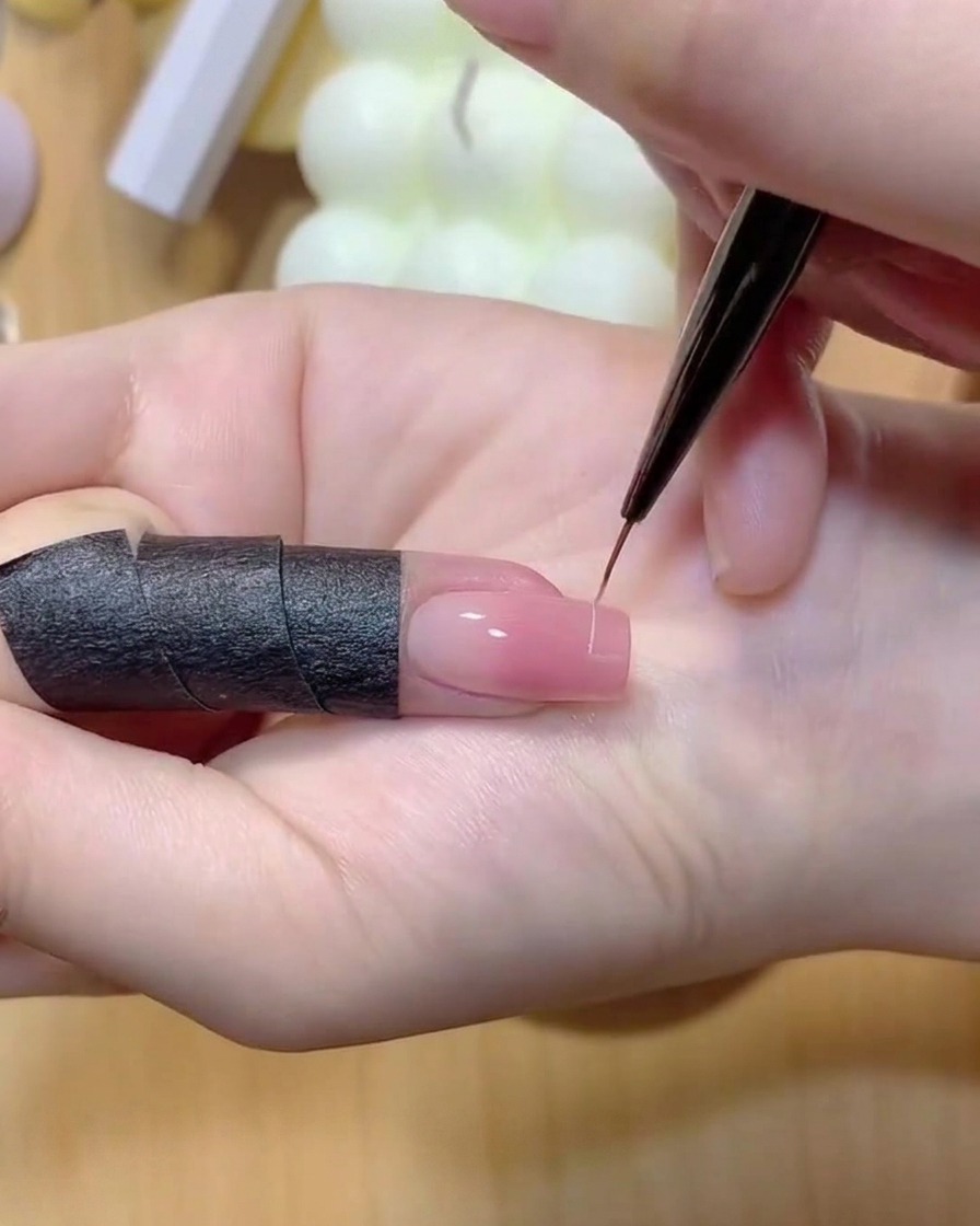 Ein feiner Liner-Pinsel zeichnet eine dünne weiße Linie über die Spitze eines rosa Farbverlaufs-Fingernagels.