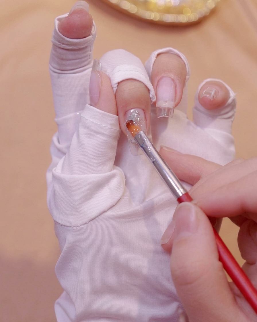 Ein feiner Pinsel trägt Silberglittergel nahe der Basis einer klaren Nagelverlängerung auf einer bemalten Hand auf.