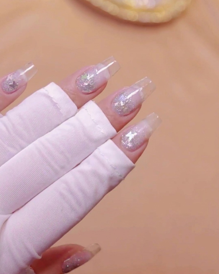 Eine Hand mit klaren Nagelverlängerungen und eingebettetem Schmetterlingsglitter unter einer UV-Lampe.