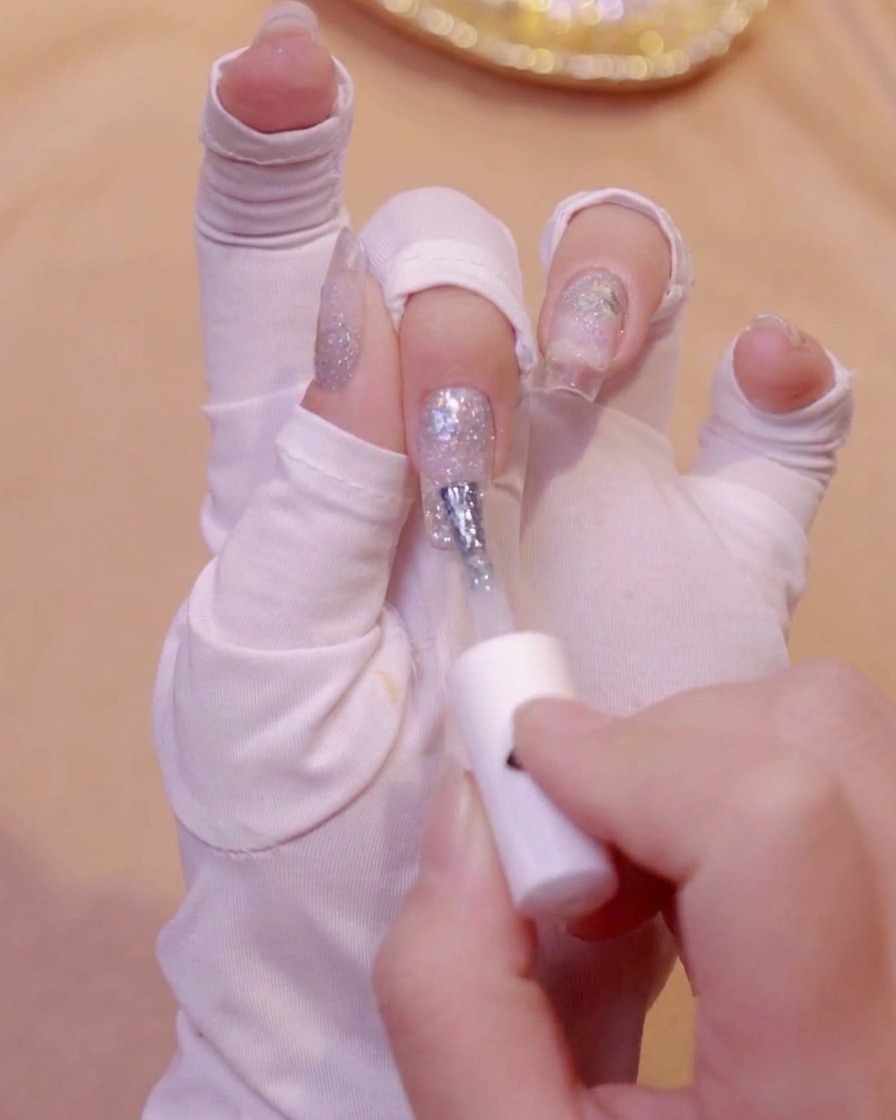 Eine weiß behandschuhte Hand trägt mit einem Pinsel milchweißes Gel-Polish auf die Spitze eines Fingernagels auf.