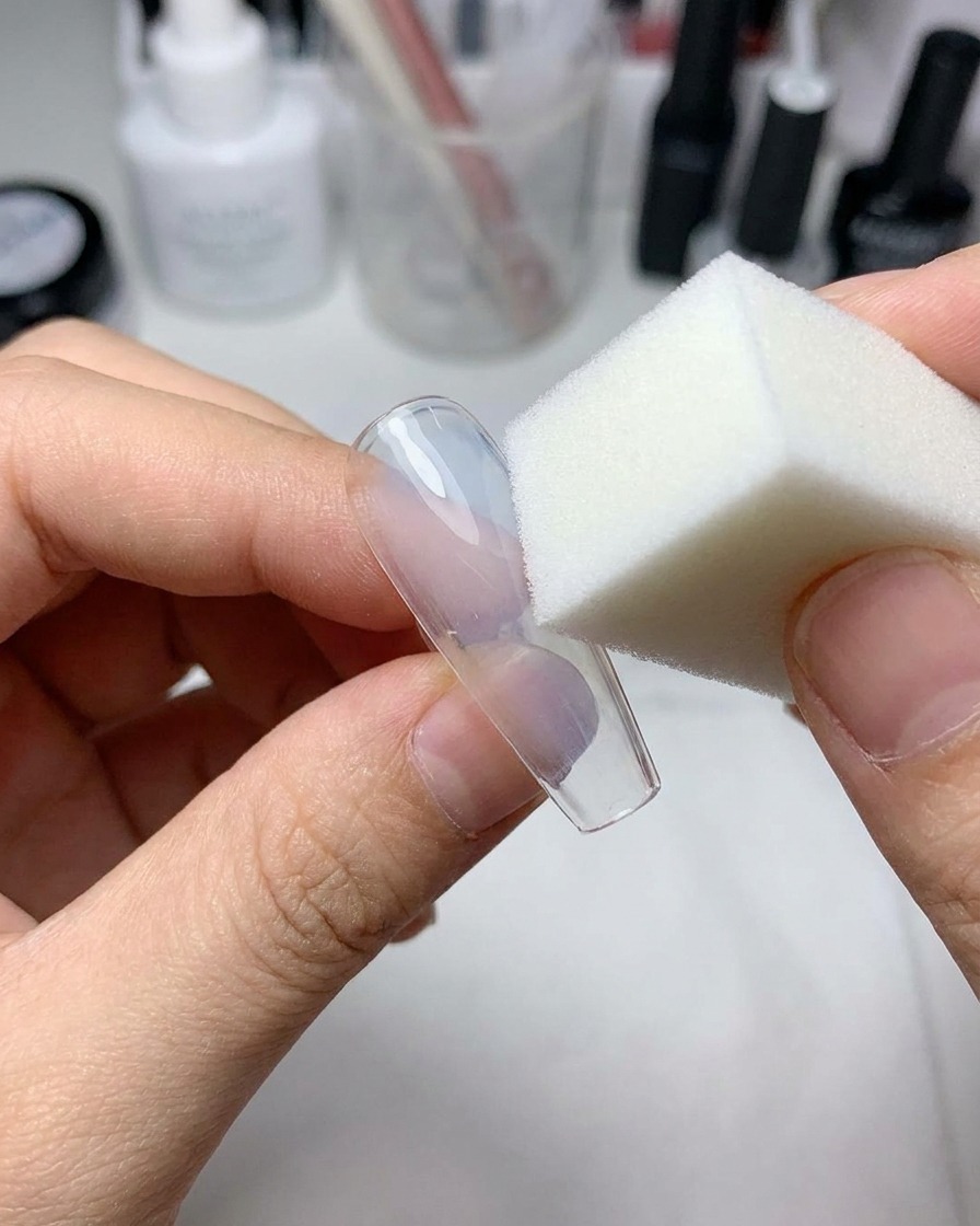 Ein kleiner Make-up-Schwamm drückt hellblaues Gel auf die Mitte eines durchsichtigen Kunstnagels.