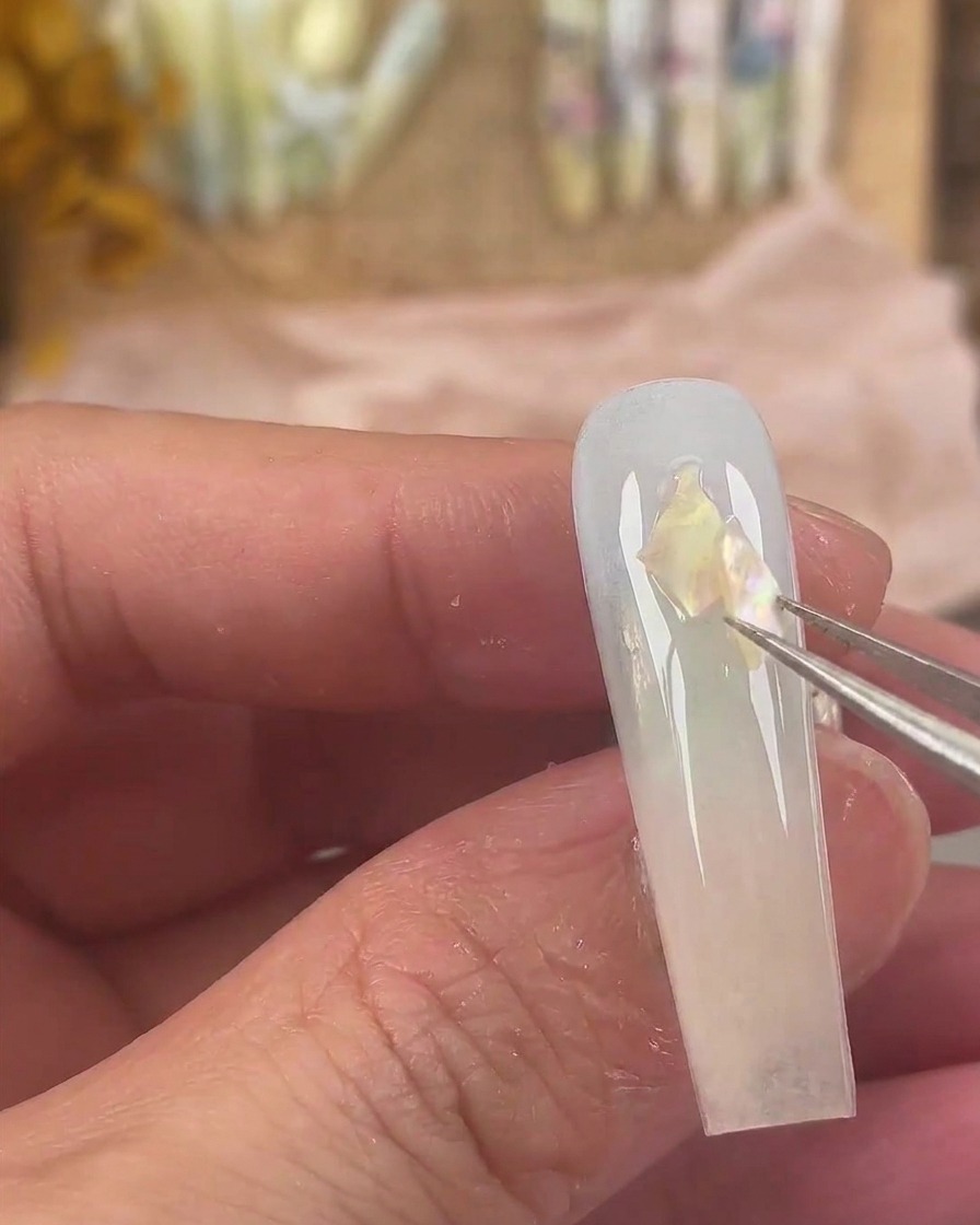 Eine feine Pinzette platziert schillernde Muschelstücke auf der nassen Geloberfläche des Nagels.