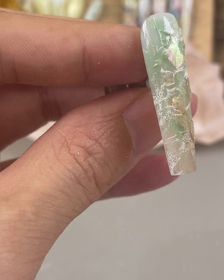 Nahaufnahme eines grünlich-klaren Nagels mit schillernden Muschelflocken und weißen Spitzenmustern, die auf die Oberfläche gestempelt wurden.