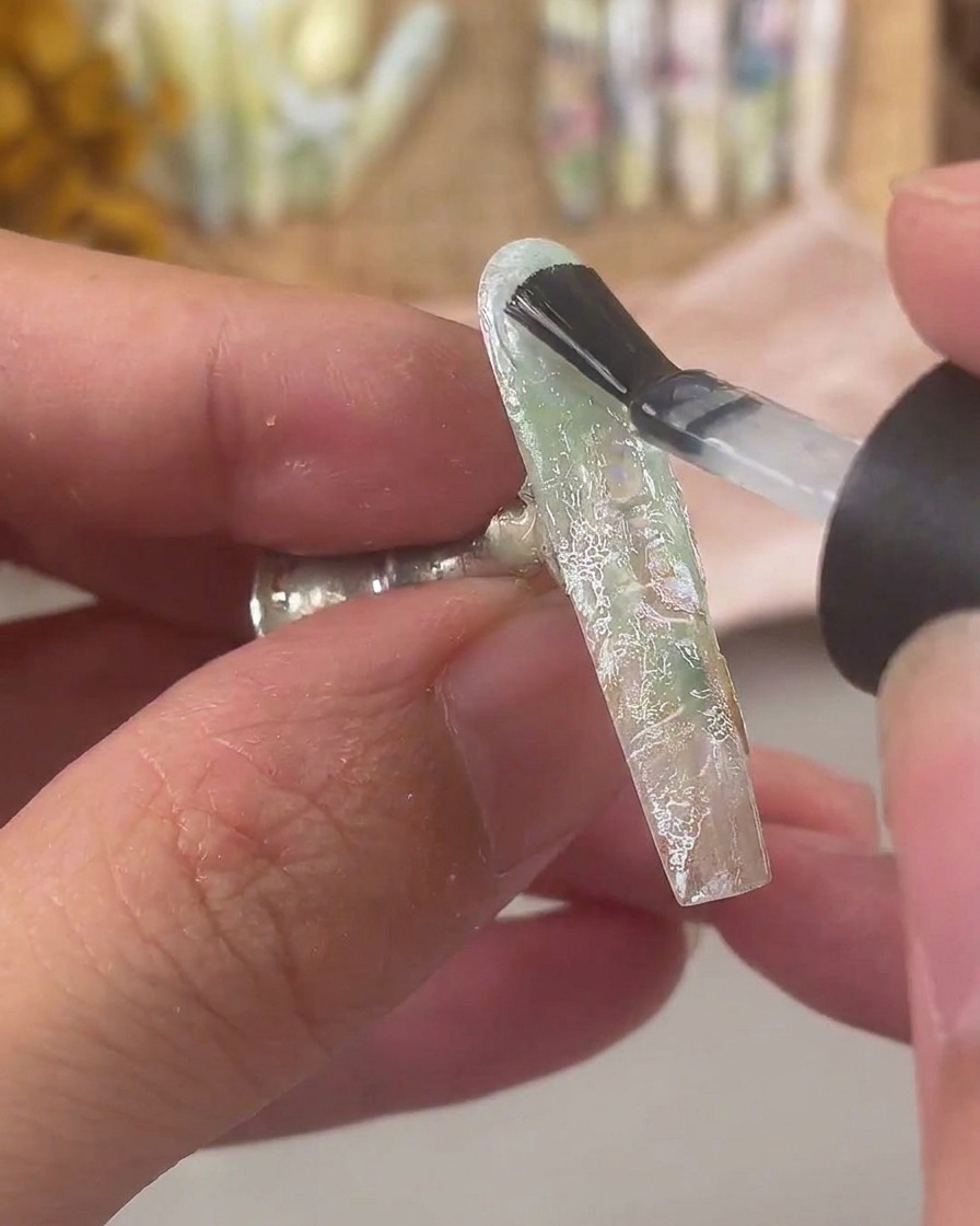 Ein Nagelpinsel trägt eine dicke Schicht klares Aufbaugel über einen mit Spitze und Muschelflocken verzierten Nagel auf.