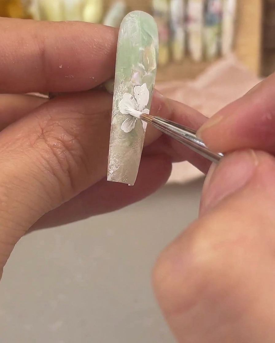 Ein feiner Detailpinsel trägt weißes Painting Gel auf, um eine große Blume auf einem mattgrünen und klaren Nagel zu zeichnen.