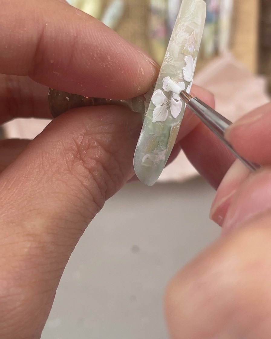 Ein feiner Detailpinsel malt drei kleine weiße Blütenblätter entlang der unteren Seitenkante eines verzierten Nagels.