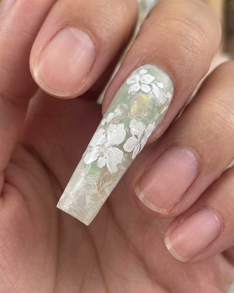 Nahaufnahme eines langen, modellierten Nagels mit fertigen weißen Gelblumen und winzigen Blättern auf einem grünen Hintergrund mit Muschelflocken.