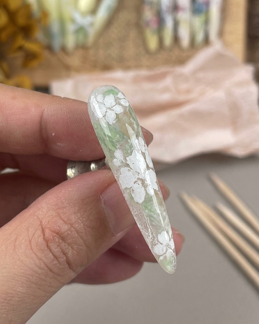 Ein langer, matt lackierter Nagel mit weißem Blumendesign, der auf einem magnetischen Nagelständer gehalten wird.