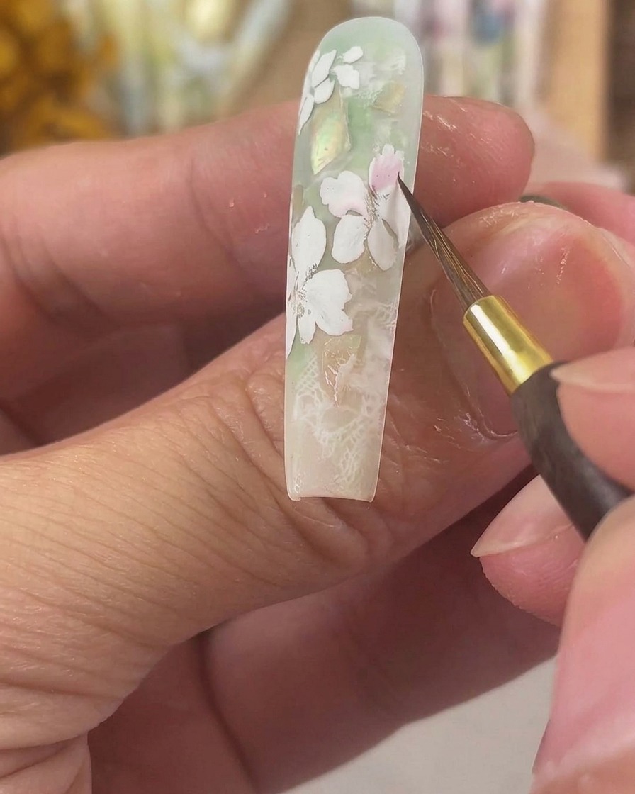 Ein feiner Detailpinsel trägt zartes rosa Aquarellpigment auf die matten weißen Gelblüten auf einem langen, modellierten Nagel auf.