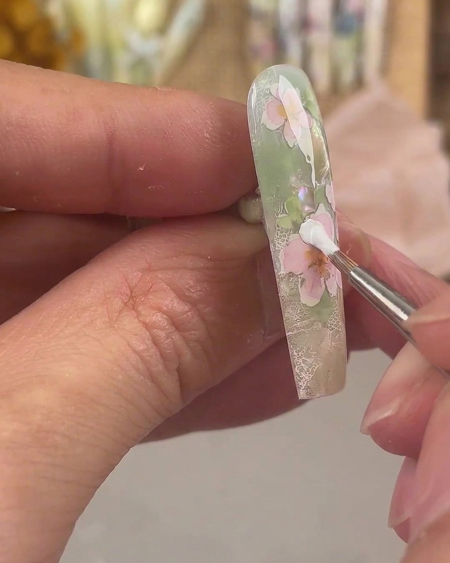 Ein feiner Liner-Pinsel trägt weißes Painting Gel auf, um eine zusätzliche Schicht Blütenblätter auf einem rosa floralen Nageldesign zu formen.