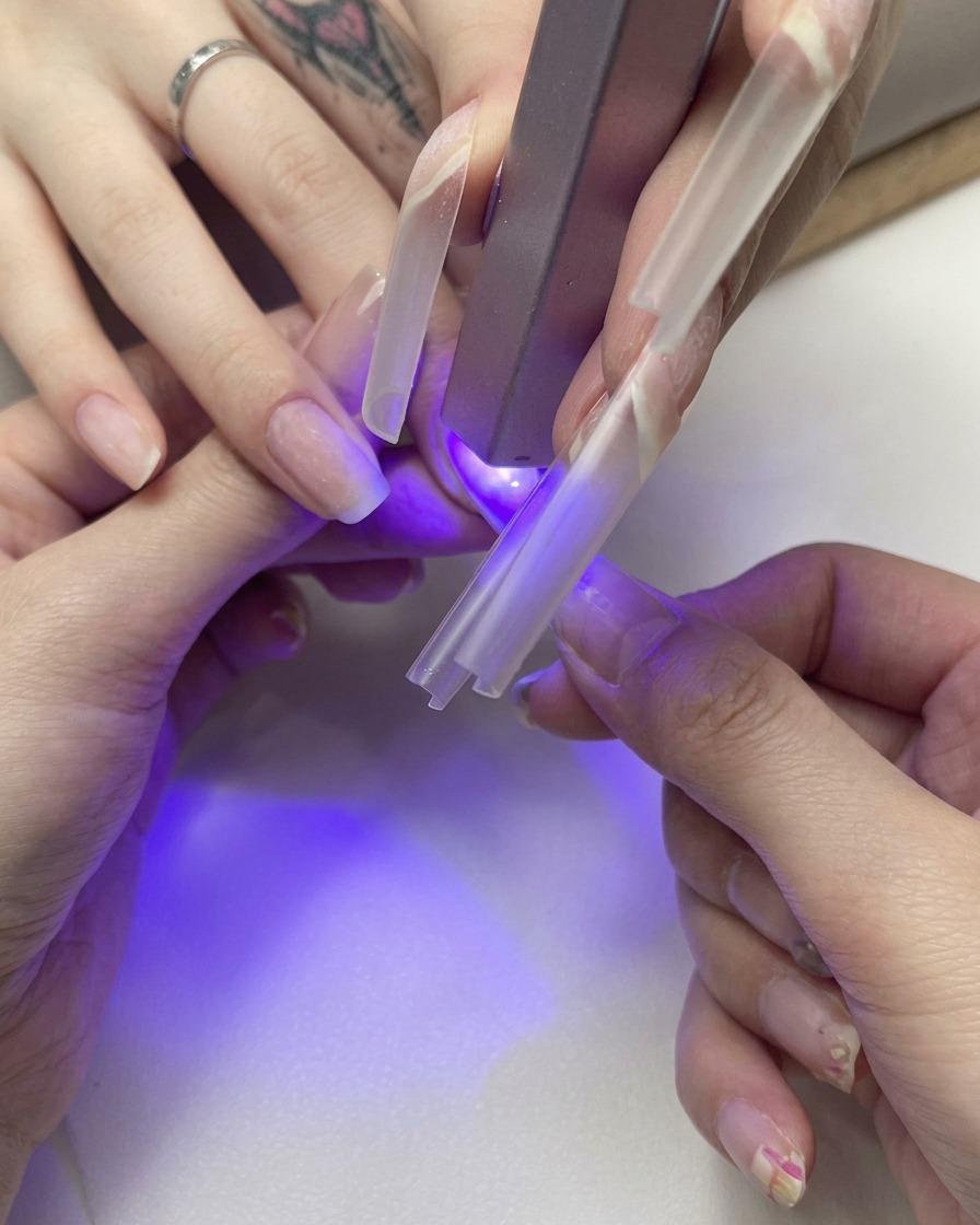 Eine Nahaufnahme einer Hand, die einen klaren Nagel-Tip auf einen Naturnagel drückt, während er unter einer kleinen violetten UV-Lampe aushärtet.
