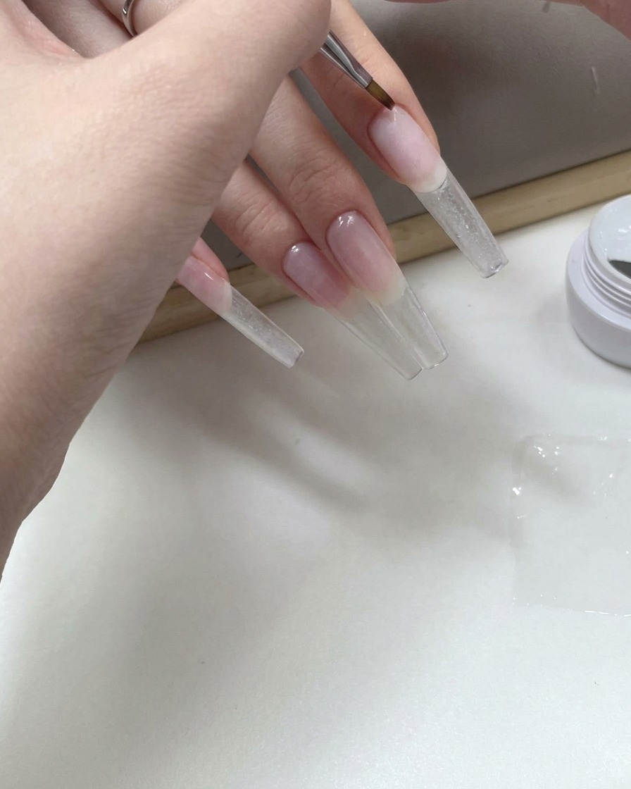 Ein feiner Nailart-Pinsel, der klares Gel auf die Unterseite eines langen transparenten Nagel-Tips aufträgt.