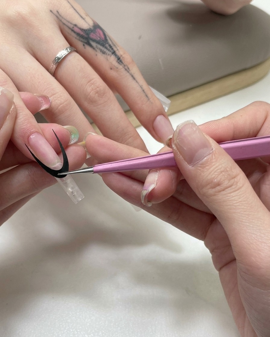 Eine rosa Pinzette bringt einen schwarzen gebogenen French-Tip Leitaufkleber auf einer klaren Nagelverlängerung an.