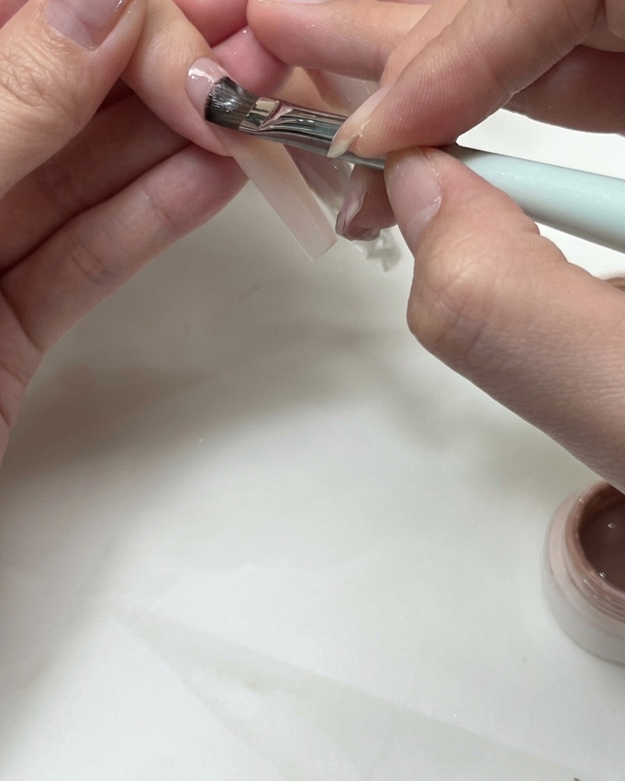 Ein flacher Pinsel trägt Nude-Gel auf, um eine French-Smile-Line auf einem Fingernagel zu formen.
