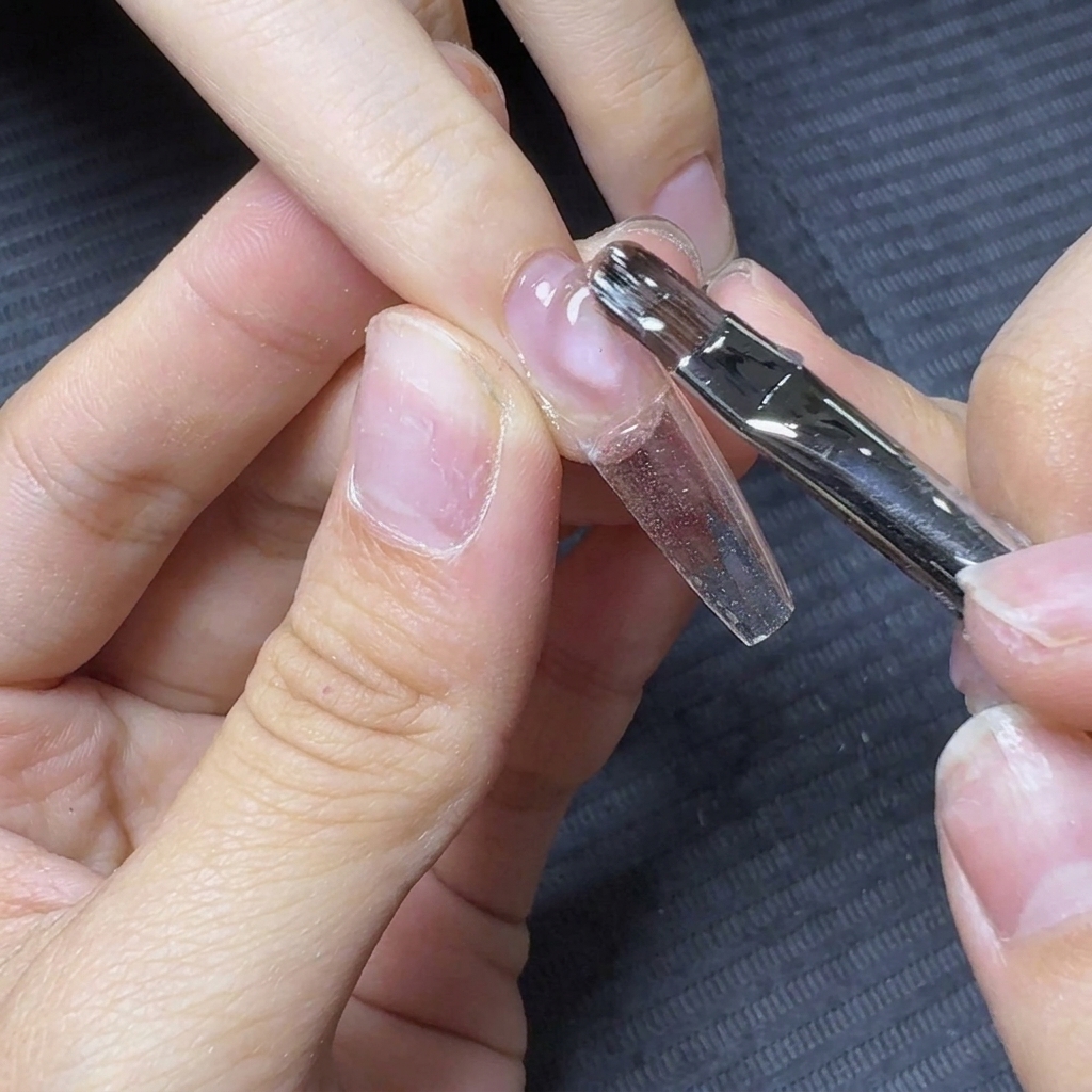 Ein Nagelpinsel zieht das klare Aufbaugel über die Länge einer klaren Nagelverlängerung.