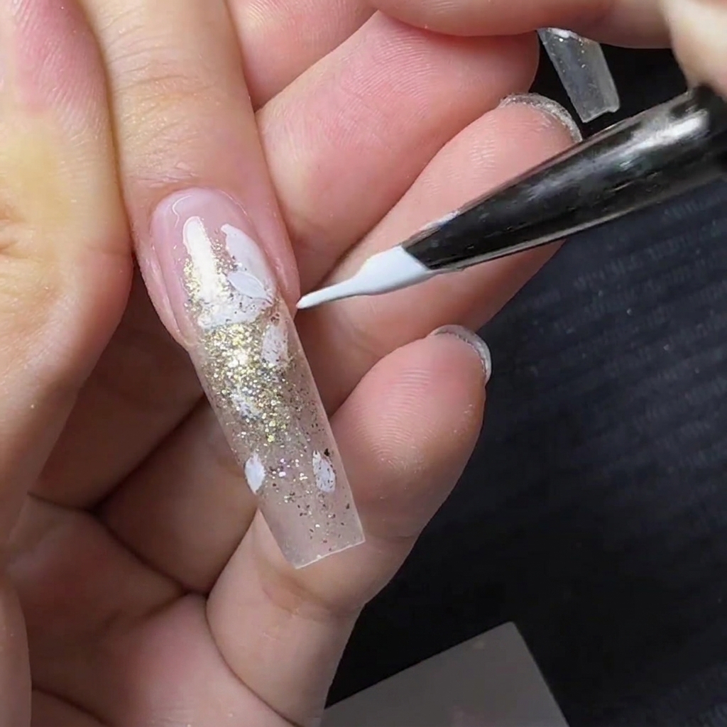 Ein feiner Liner-Pinsel malt kleine weiße Blütenblätter über einen klaren Nagel mit Goldglitter.