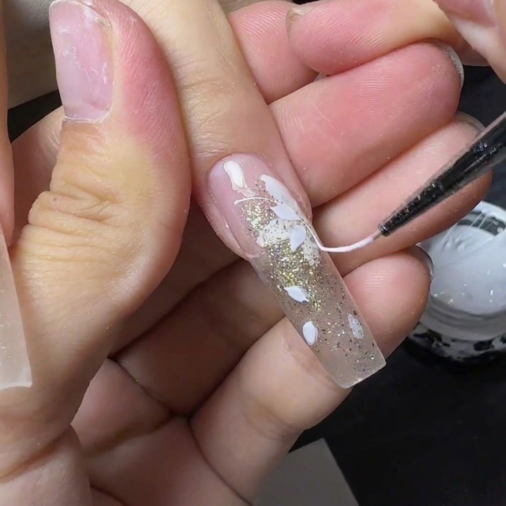 Ein dünnes Werkzeug zieht fadenartiges weißes Spider-Gel über einen mit weißen Blumen verzierten Nagellack.