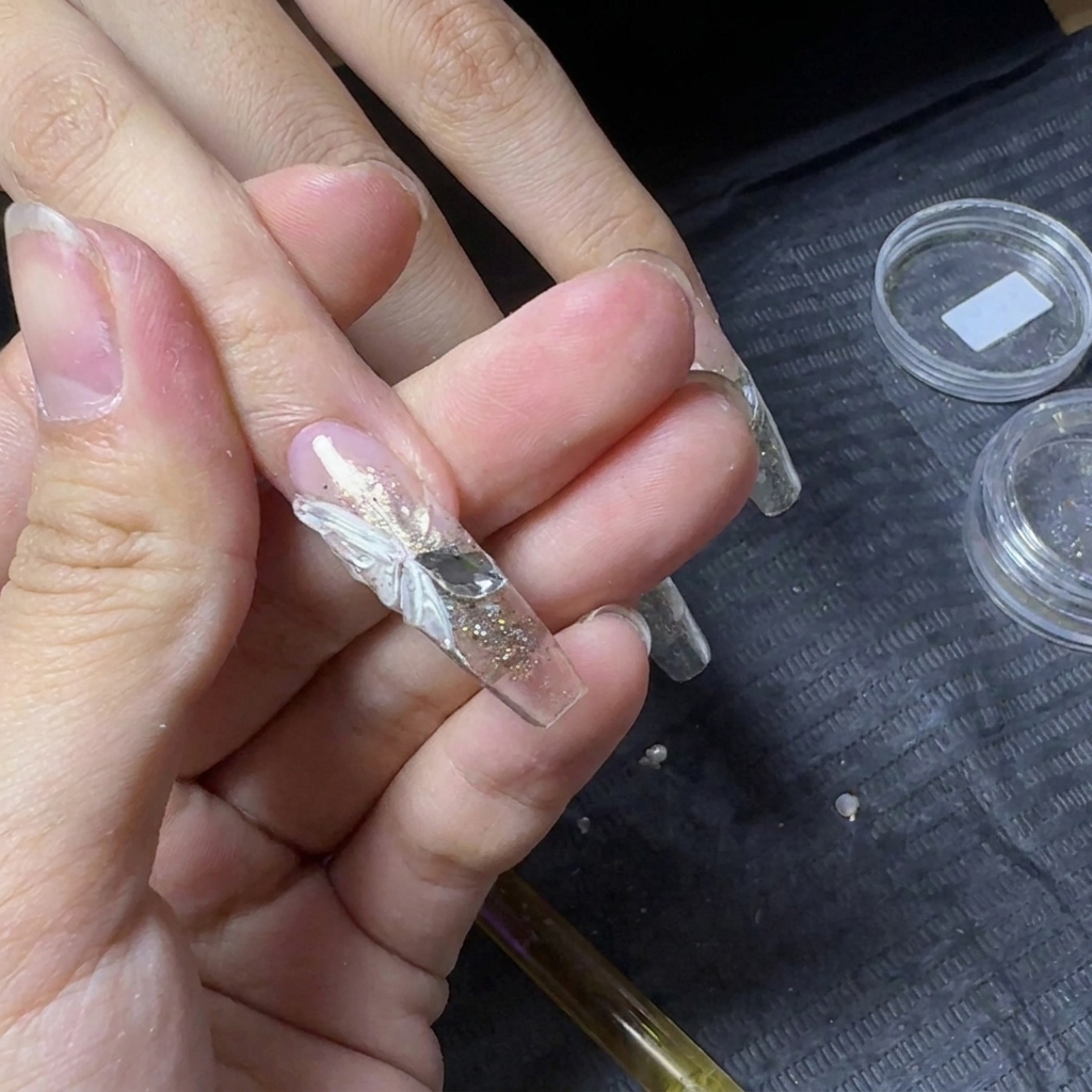 Nahaufnahme einer klaren Nagelverlängerung mit einem weißen 3D-Schmetterlingsflügel und zwei Flachsteinen.