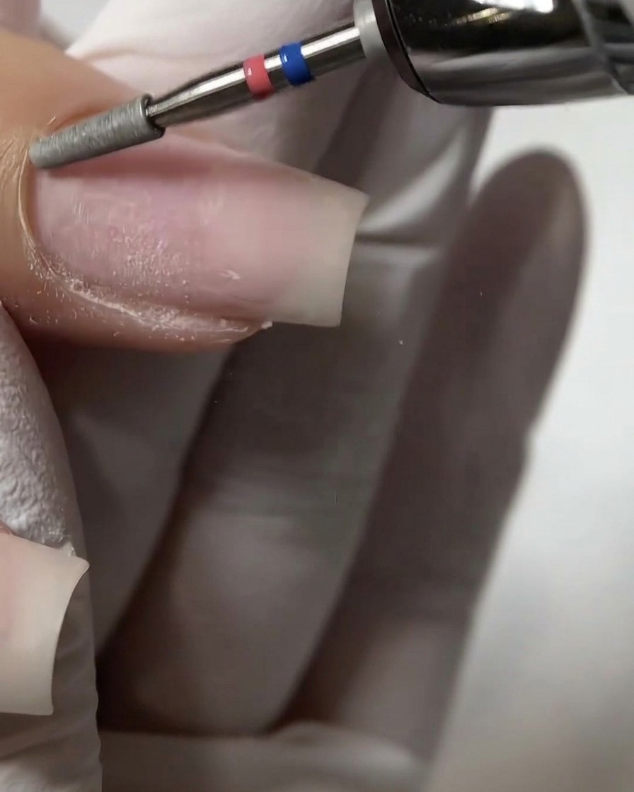 Eine Nahaufnahme eines flachen Zylinder-Fräseraufsatzes, der sanft die proximale Nagelhautfalte an einem Naturnagel exfoliert und anhebt.