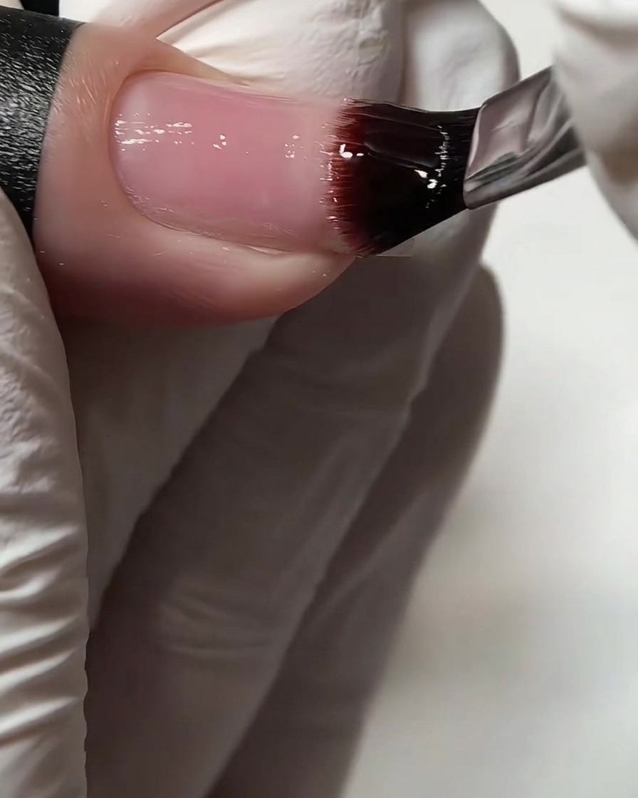 Eine Nahaufnahme eines flachen Gelpinsels, der einen klaren Base Coat auf einen Naturnagel aufträgt.