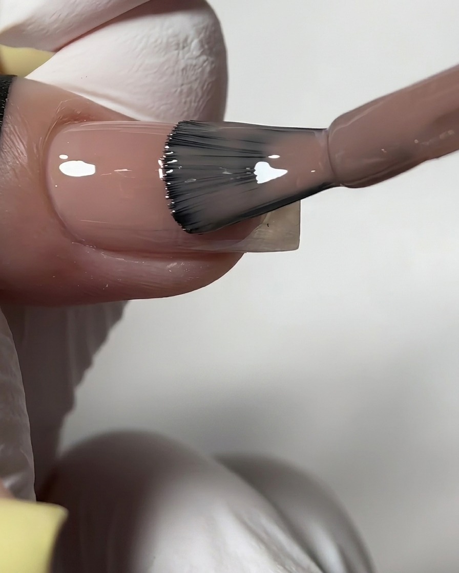 Ein Flaschenpinsel trägt einen transparenten, glänzenden Nude-Gelack auf einen vorbereiteten Fingernagel auf.