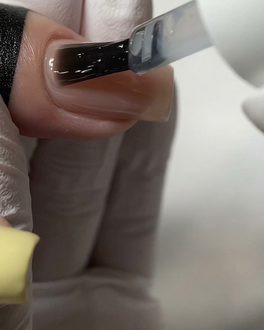 Ein Pinsel trägt einen klaren Top Coat über einen nudefarbenen Acrylnagel auf.