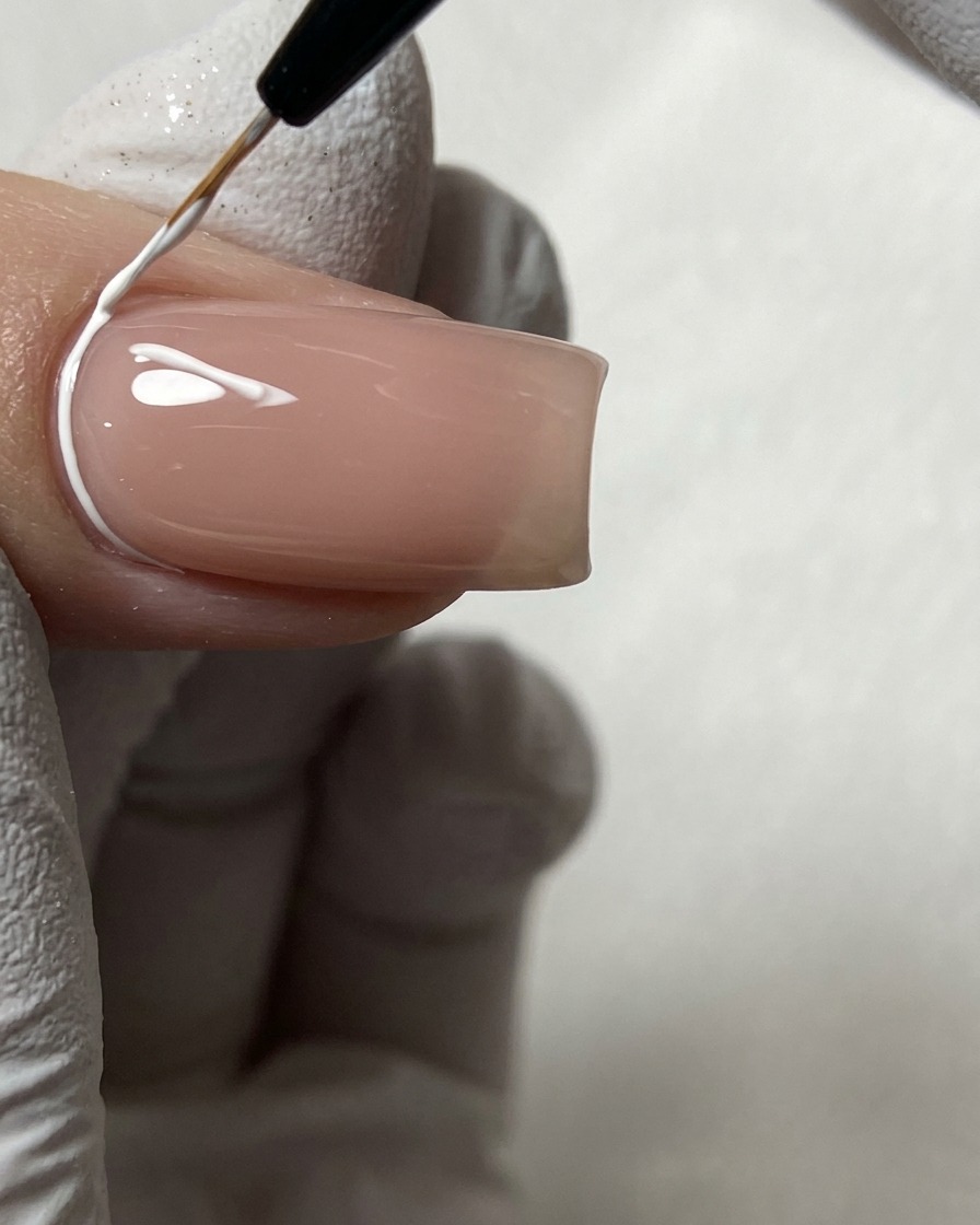 Ein feiner Nailart-Pinsel zeichnet weiße geometrische Linien nahe der Nagelhaut auf einen Nude-Nagel.