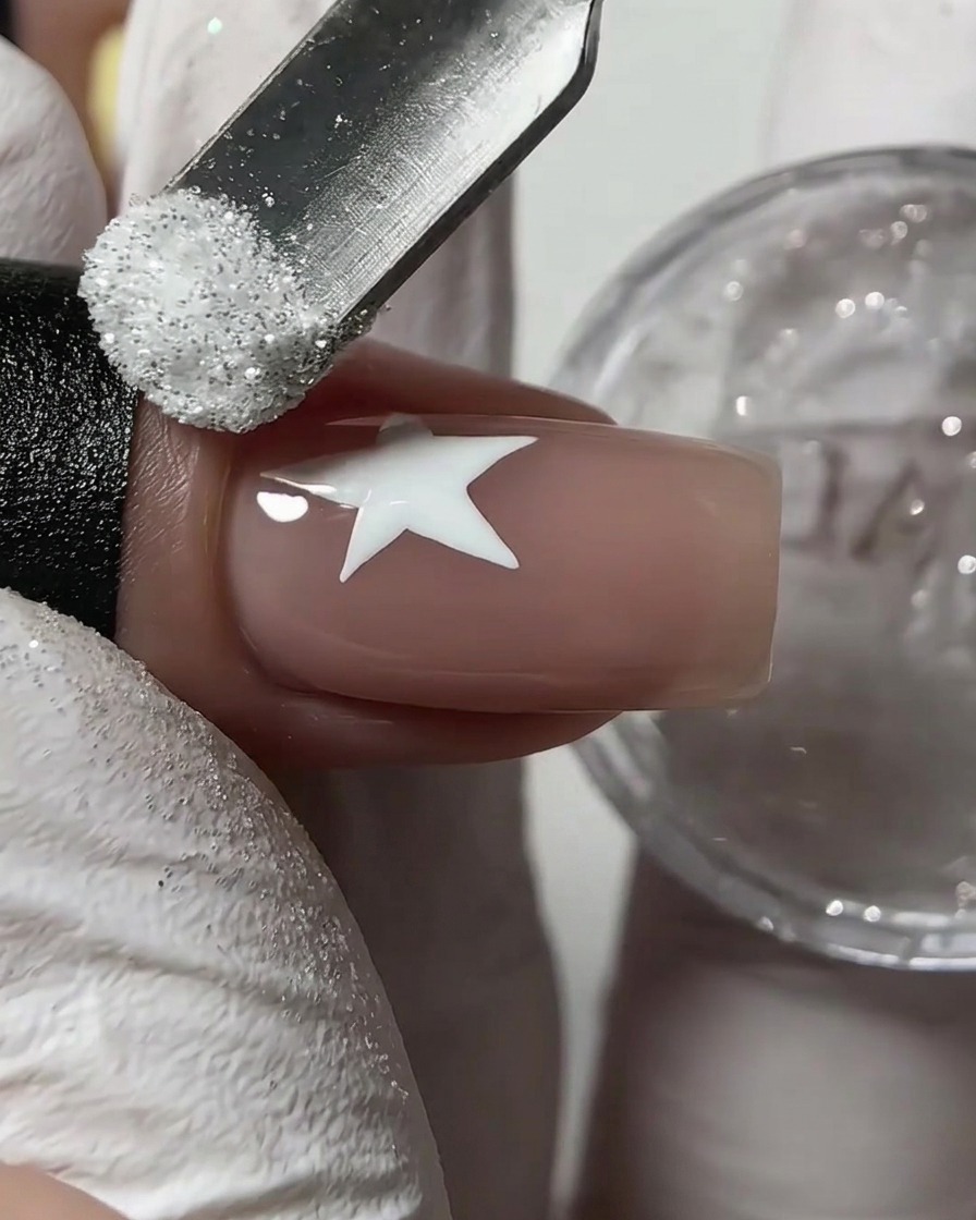 Ein Nagelhautschieber aus Metall streut weißen Zucker-Glitzer auf einen feuchten weißen Stern auf einem Nude-Fingernagel.