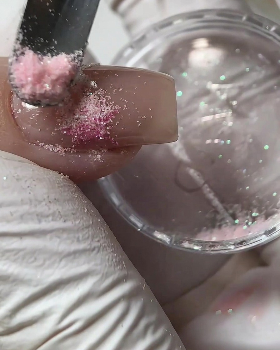 Ein kleines Werkzeug streut rosa Zuckerpulver über ein feuchtes Gel-Design auf einem Fingernagel.