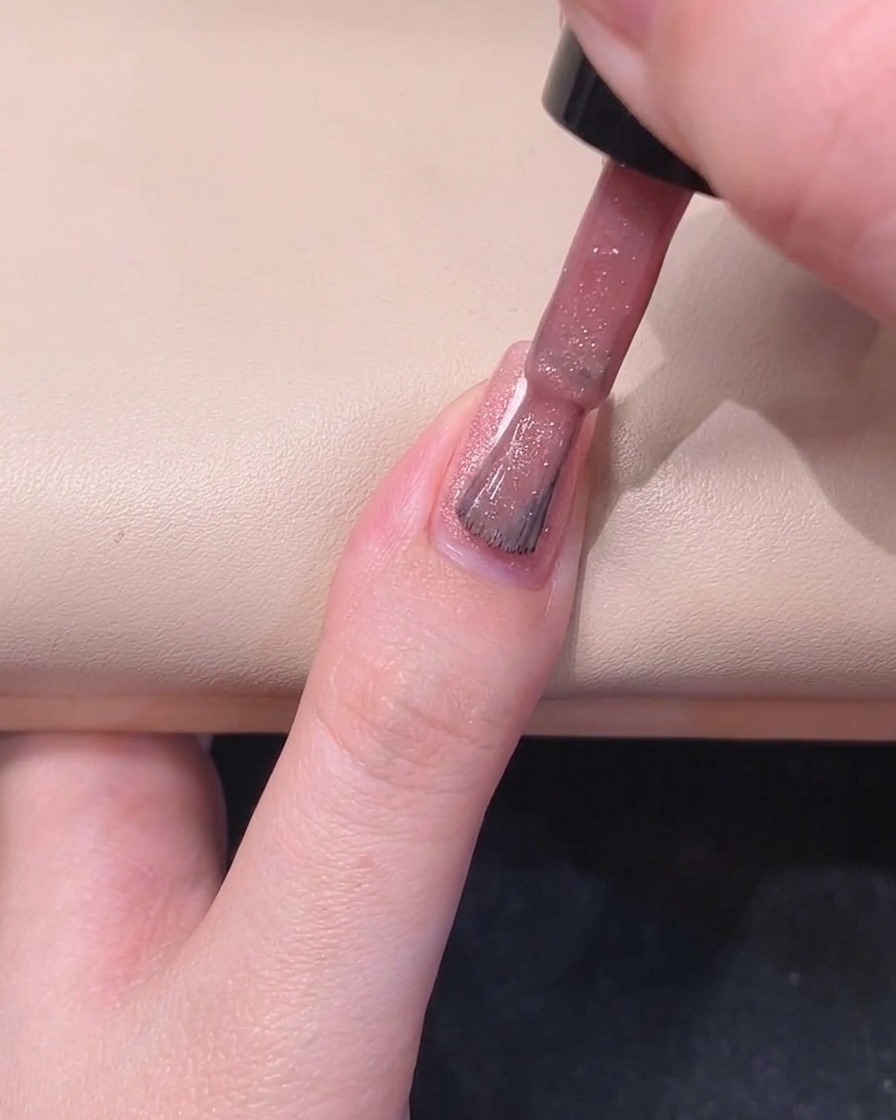 Ein Nagelpinsel bemalt die rechte Seite eines Fingernagels mit schimmerndem rosa Gelack.