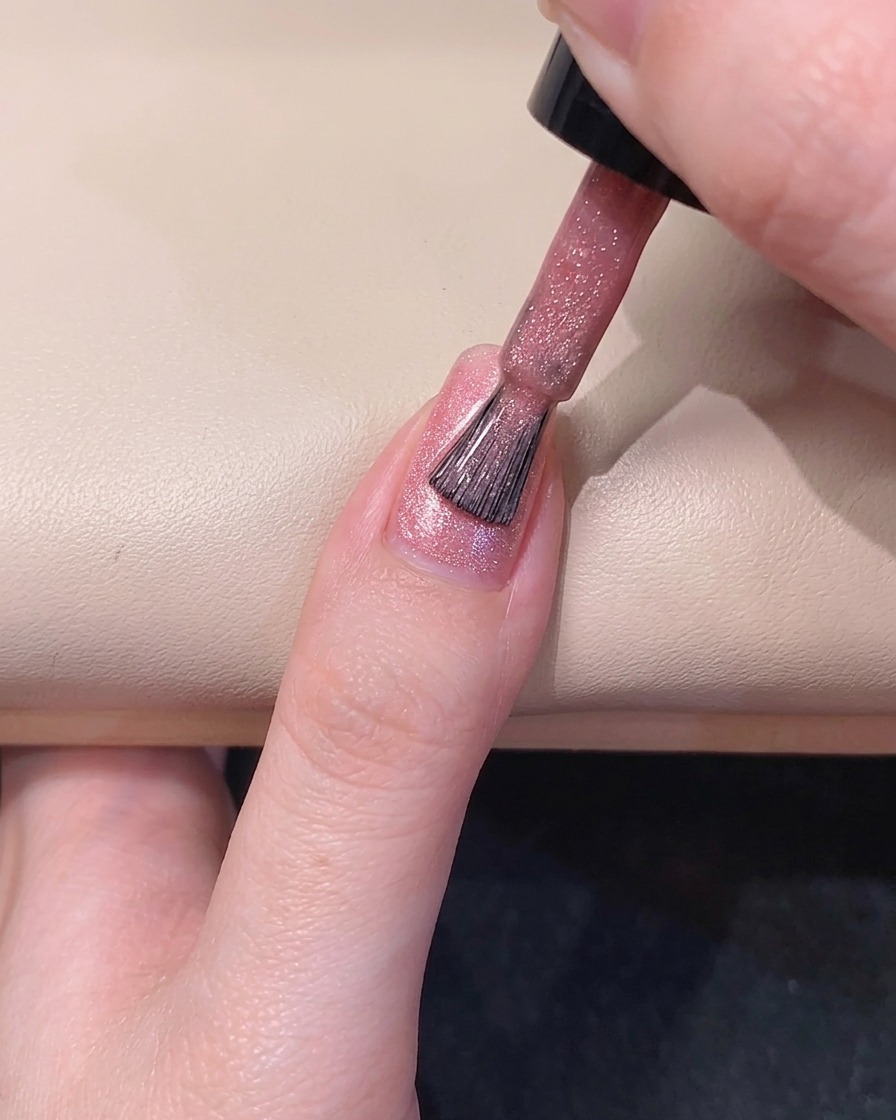 Ein Nagelpinsel verteilt zartrosa Magnet-Gelack an der linken Seite des Fingernagels.