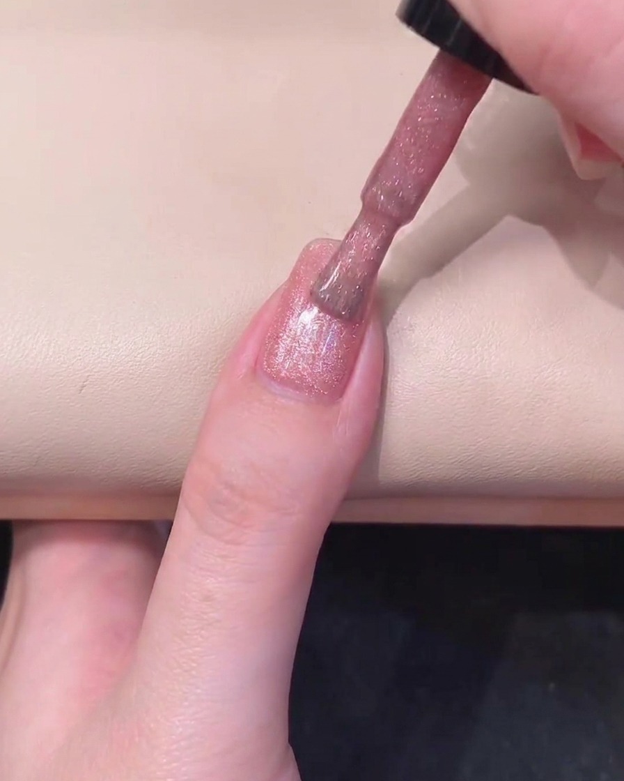 Ein schwarzer Zylindermagnet wird parallel zur rechten Seite eines rosa gellackierten Nagels gehalten, um den Schimmer zu aktivieren.