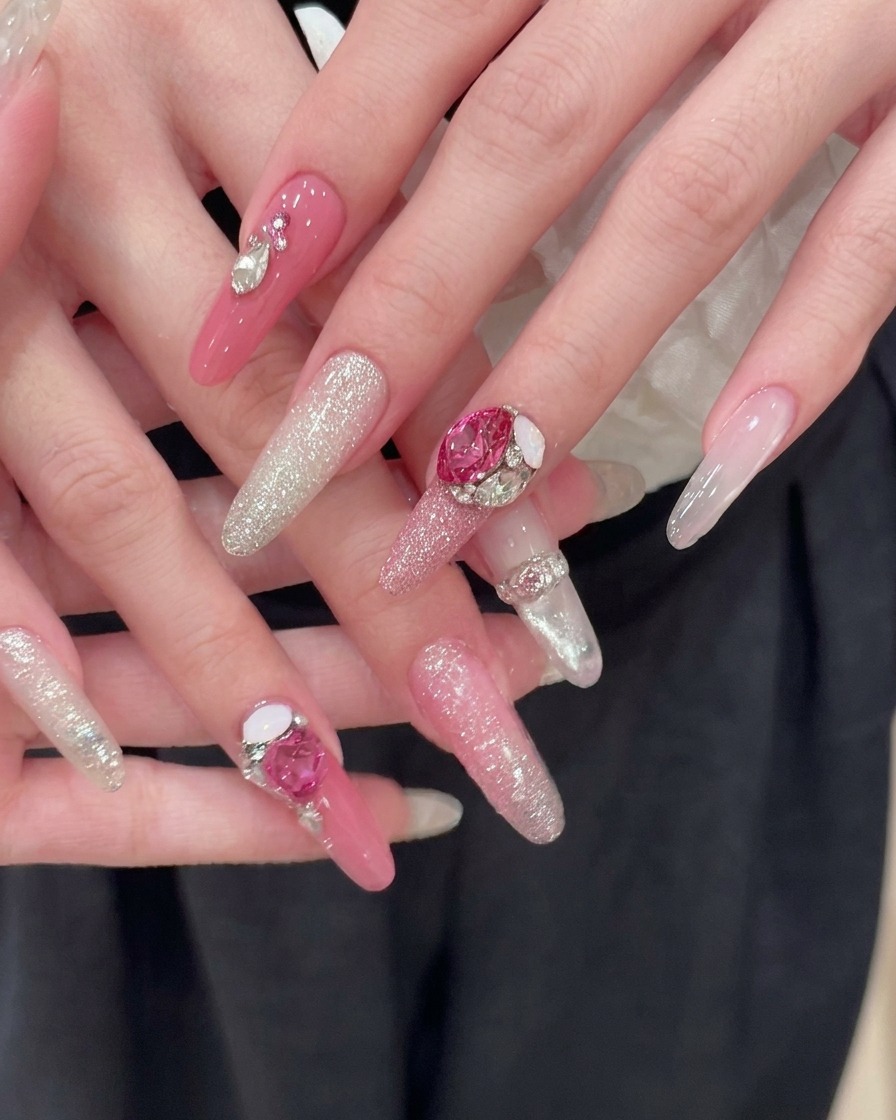 Zwei perfekt manikürte Hände ruhen vor einem dunklen Stoffhintergrund und zeigen fertige lange rosa Mandelnägel mit Silberglitter-Verläufen und großen rosa Herz-Strass-Akzenten.